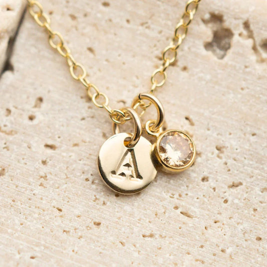 Gold initial 'A' pendant necklace with a round light brown (champagne-colored) birthstone charm on a delicate chain, displayed on a textured light stone surface