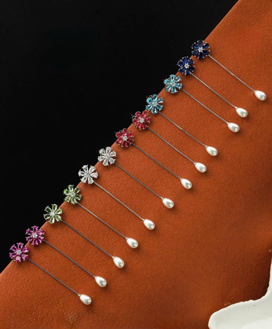 Row of colorful gemstone flower pins—in vibrant blues, red, clear, green, and pink—each with a delicate chain leading to a white teardrop pearl, displayed on a rust-orange textured surface.