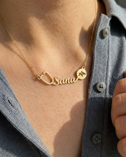 Elegant personalized medical-themed necklace with name, stethoscope, and heartbeat charm in 22K gold finish, worn with a neutral outfit in warm sunlight.