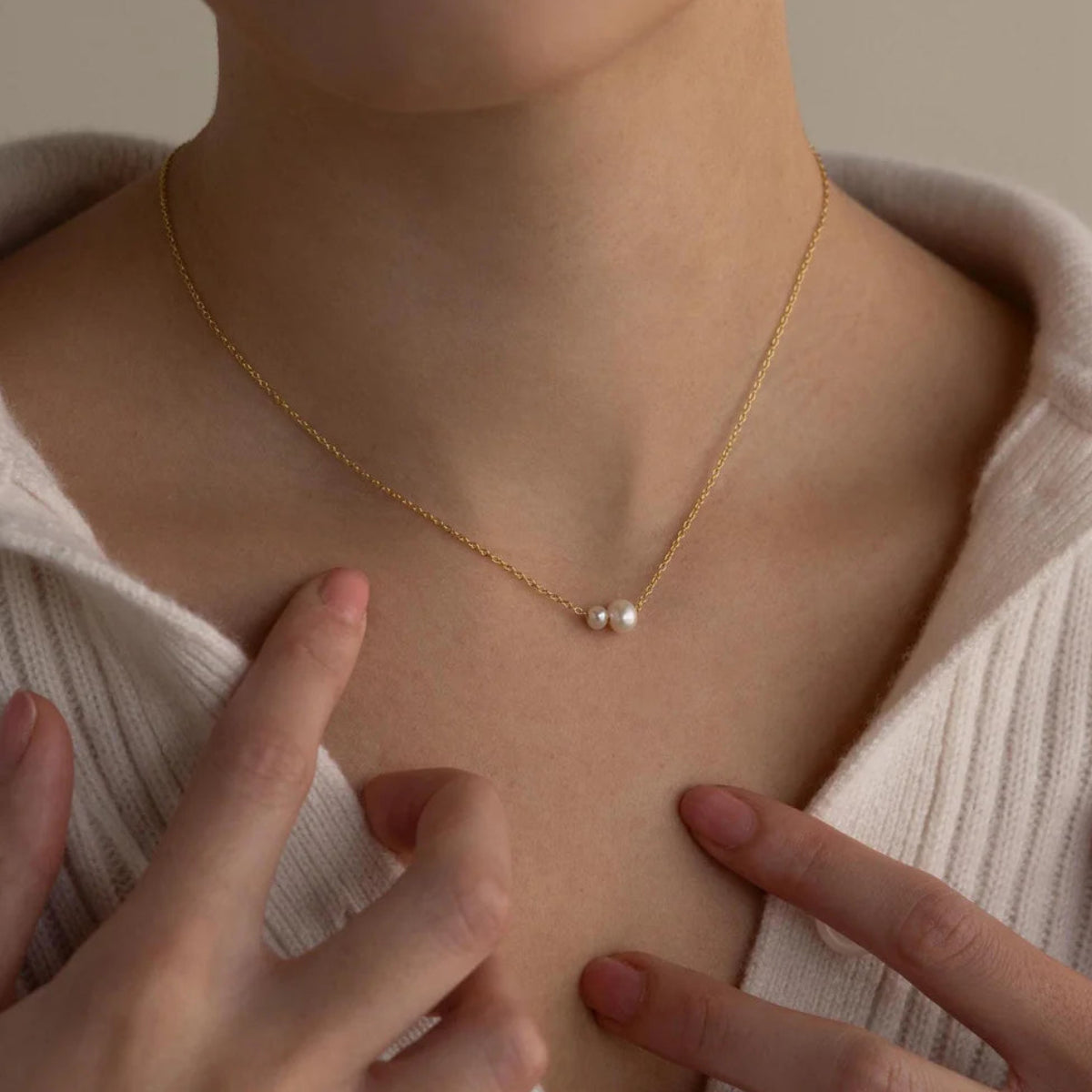 Person wearing a delicate gold necklace with small pendants, pointing to it with their fingers.