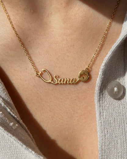 Close-up of a gold personalized name necklace featuring a stethoscope design and heartbeat symbol, worn on the neck with a soft sunlit aesthetic.