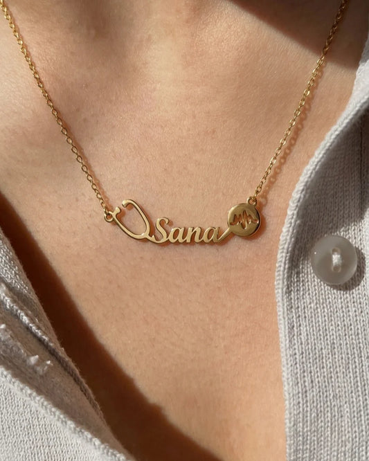 Close-up of a gold personalized name necklace featuring a stethoscope design and heartbeat symbol, worn on the neck with a soft sunlit aesthetic.