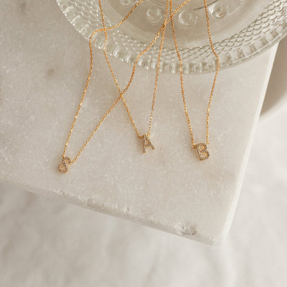 Three delicate gold initial necklaces with letters S, A, and B, displayed on a white marble block next to a clear glass dish.